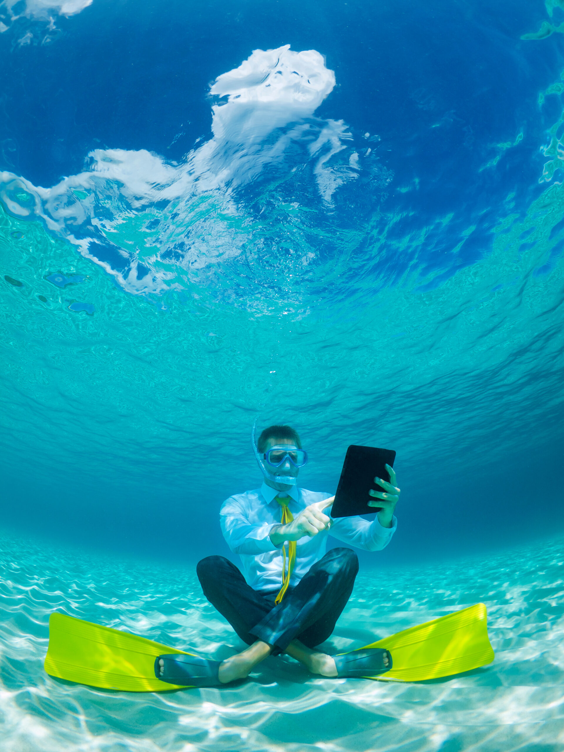 Snorkelling businessman in shirt and tie and matching fins using a tablet computer sitting underwater in tropical turquoise sea